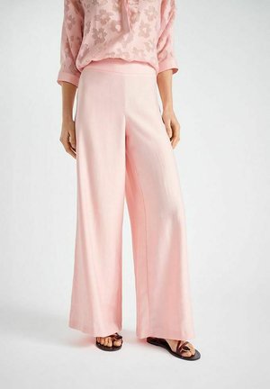 Vrouw draagt lichtroze broek met wijde pijpen, een doorzichtige blouse met bloemenpatroon, en zwarte platte sandalen met open tenen, staand tegen een witte achtergrond.