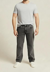 Un homme est debout portant un t-shirt gris clair à manches courtes, un jean gris foncé et des baskets blanches sur un fond beige uni.