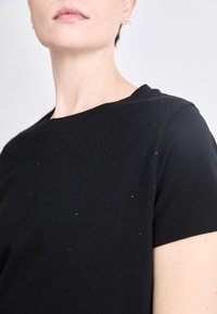 Melns īsās piedurknes t-krekls ar apaļu kakla izgriezumu, ar smalkām spīdošām rotājumām, kas vienmērīgi sadalītas pa priekšpusi.