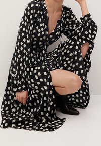 Vestido negro con patrón cuadrado blanco, mangas largas y diseño fluido. La tela parece ligera con una textura suave. Botas negras visibles.