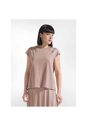Vrouw met stijl zwart haar die een loszittend, beige topje met korte mouwen en een bijpassende rok draagt, staand tegen een witte achtergrond.