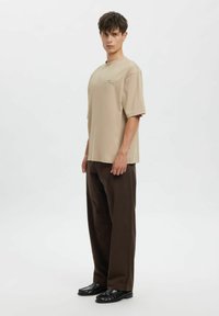 Béžové oversized tričko s krátkými rukávy, měkkou texturou, ve spojení s hnědými širokými kalhotami a černými mokasínami. Minimalistický design s detailem značky.