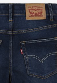 Jeans in denim blu scuro con cuciture gialle, caratterizzati da un'etichetta in pelle in vita e un piccolo etichetta rossa sulla tasca posteriore.