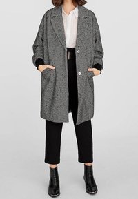 Een houndstooth-gepatenteerde jas in zwart en wit, met een losse pasvorm, oversized kraag, zijzakken en opgerolde mouwen, gecombineerd met zwarte broeken.