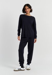 Marineblauer Strickpullover mit lockerer Passform, kombiniert mit passenden Jogginghosen und weißen Sneakers mit schwarzen Akzenten. Glatte Textur.