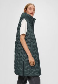 Gestepptes türkisfarbenes Weste mit hohem Kragen, Seitentaschen und Reißverschluss an den Seiten. Glatte Textur mit geometrischem Muster überall.