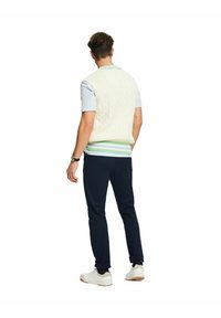 Homme portant un gilet en tricot blanc avec des rayures vertes et bleues, une chemise bleu clair, un pantalon foncé, des baskets blanches et une montre-bracelet, vu de dos.