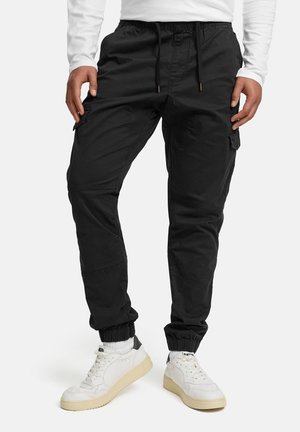 Homme portant un pantalon de jogging cargo noir avec des poignets élastiques, des baskets blanches et une chemise à manches longues blanche, debout devant un fond blanc.