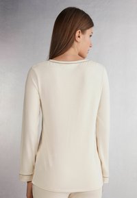 Langsleeve beige top van zacht materiaal met een ontspannen pasvorm, ronde halslijn en subtiele zijsplitten, met een gladde textuur en minimaal ontwerp.
