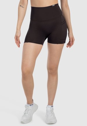 Kvinde iført sorte atletikshorts med høj talje og sidelommer samt hvide sneakers, stående mod en ensfarvet lys baggrund.