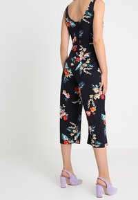 Svart blommig jumpsuit med färgglada blommor, åtsittande livstycke, breda ben, längd till mitten av vaden och en låg rygg. Matchad med ljuslila klackar.