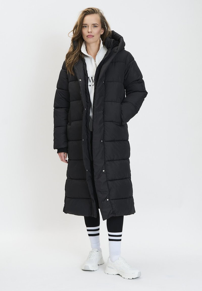 Abrigo de plumas negro con capucha, diseño acolchado, botones de presión y un ajuste hasta la rodilla, combinado con una sudadera blanca con cremallera y leggings negros.