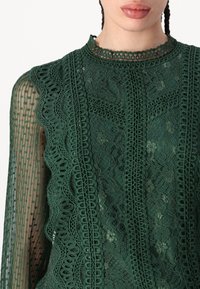 Top in pizzo verde con motivi floreali intricati, dettagli testurizzati e un colletto alto con finiture decorative. Presenta maniche lunghe in tessuto trasparente.