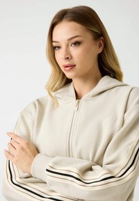 Beżowa bluza z zamkiem, o prążkowanej teksturze, z kapturem, haftowanym logo oraz paskami w kolorze czarnym, kremowym i rdzy wzdłuż rękawów.