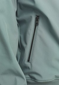 Zeleni zatvarač na mekoj, teksturiranoj tkanini s glatkom površinom; uključuje crne metalne dijelove zatvarača i detalje šivanja.