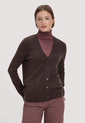 Frau mit glattem Haar, die eine dunkelbraune Strickjacke über einem mauvefarbenen Rollkragenpullover und mauvefarbenen Hosen vor einem schlichten Hintergrund trägt.