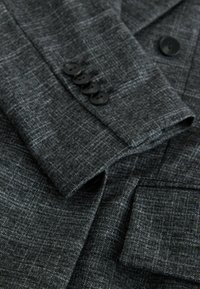 Detalle de un blazer gris oscuro con una tela texturizada, cuatro botones negros en el puño y un bolsillo de parche con un sutil patrón de cuadros.