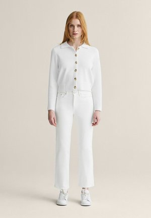 Kvinde med rødt hår iført en hvid knappet cardigan, hvide højtaljede lige bukser og hvide sneakers, stående mod en beige baggrund.