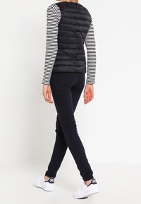 Svart pufferväst med quiltat mönster, bärs över en svart-vit randig långärmad skjorta. Kombineras med marinblå byxor och vita sneakers.