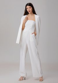 Combinaison blanche avec un corsage ajusté, un pantalon à jambes larges et des poches latérales. Accompagnée d'un blazer blanc sur mesure. Tissu lisse et brillant.