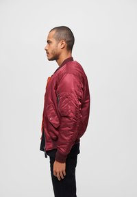 Veste bomber bordeaux avec finition brillante, poignets côtelés, fermeture éclair et poches latérales ; dotée d'une doublure orange et d'une poche sur la manche.