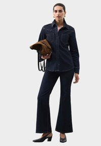 Mørk denimskjorte med to brystlommer, parret med vide mørke denimbukser. Holder en brun taske, har sorte højhælede sko på.