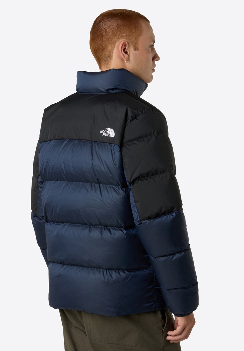 Marineblaue Steppjacke mit schwarzen Akzenten. Sie hat einen hohen Kragen, ein horizontal gestepptes Design und das Logo von The North Face auf der linken Schulter.
