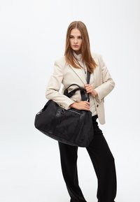 En kvinna med långt hår poserar självsäkert i en beige kavaj, håller en stilren svart väska, mot en enkel vit bakgrund.