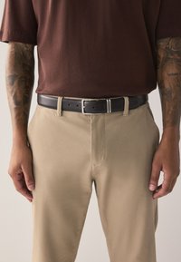 Black leather belt with silver buckle worn over beige pants and a brown short-sleeve shirt; visible tattoo detail on arms.