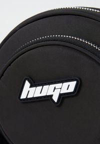 Zaino in nylon nero con design circolare, hardware lucido e un logo bianco prominente. Superficie testurizzata con dettagli con zip.