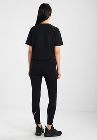 Frau mit langen dunklen Haaren, die steht und ein schwarzes, kurzes T-Shirt, schwarze Leggings mit einem kleinen pinken Logo und schwarze Turnschuhe trägt, mit dem Rücken zur Kamera.