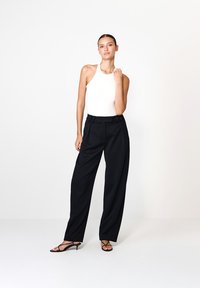 Biała bezrękawnikowa bodysuit w połączeniu z czarnymi spodniami o wysokim stanie, z dopasowanym krojem i plisami, uzupełniona czarnymi sandałami na paskach.