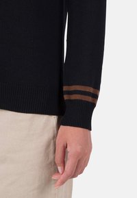 Schwarzer Strickpullover mit gerippten Bündchen und braunen Streifen. Eine sichtbare Hand ruht auf einem hellen, strukturierten Stoff.
