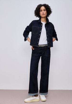 Femme aux cheveux bouclés portant une veste en jean foncé et un pantalon assorti, une chemise blanche, une ceinture imprimée léopard et des baskets blanches, debout avec les mains sur les hanches.