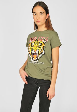 Olijfgroene katoen T-shirt met een tijgerafbeelding en "TOP GUN" in rood en wit. Korte mouwen, relaxte pasvorm. Gecombineerd met zwarte jeans.