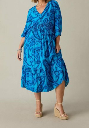 Vrouw die een blauwe midi-jurk draagt met donkere paisleymotieven, driekwart mouwen en beige sleehakken, staand tegen een neutrale achtergrond.