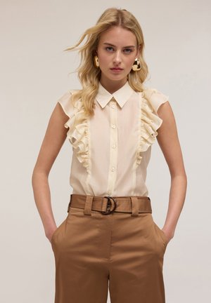 Crème-kleurige blouse met ruches langs de voorkant, korte mouwen en knoopsluiting, gecombineerd met bruine high-waisted broek.