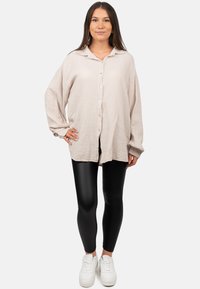 Beige overhemd met lange mouwen en knopen in een gestructureerde stof, gecombineerd met zwarte strakke legging en witte sneakers, wat een casual outfit oplevert.
