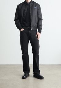 Schwarze Bomberjacke mit Reißverschluss, gerippten Bündchen, getragen über einem schwarzen Rollkragenpullover. Kombiniert mit dunklen Jeans und schwarzen Schuhen. Einfacher, monochromer Fokus.