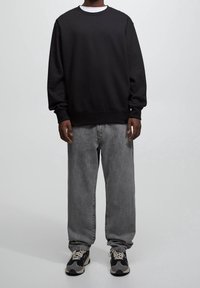 Svart sweatshirt, lös passform, rund halsringning, bärs över en vit skjorta. Kombinerad med grå raka jeans och svarta sneakers med detaljer.