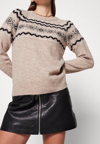 Pullover beige a maglia con motivo geometrico nero, scollatura rotonda e orlo a coste, abbinato a una mini gonna nera in pelle con zip.
