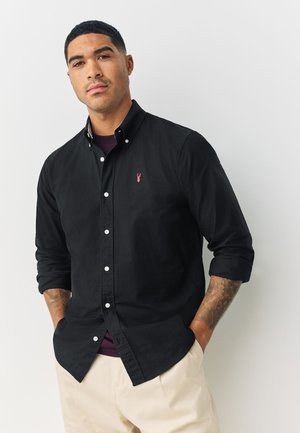 Schwarzes Button-down-Hemd mit Kragen und pinkem Hummer-Logo. Verfügt über geknöpfte Manschetten und eine leicht taillierte Passform, kombiniert mit hellbeigen Hosen.