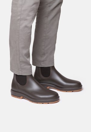 Hnedé nepremokavé členkové boots so elastickými bočnými panelmi a textúrovanou gumovou podrážkou, skombinované so svetlo šedými nohavicami.