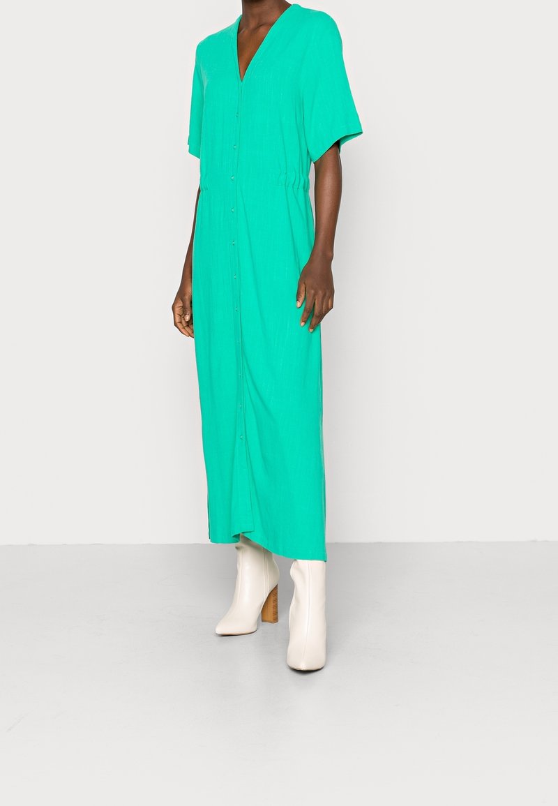 Turkoosi maksimekko V-pääntiellä ja lyhyillä hihoilla, jossa on nappikiinnitys edessä ja elastinen vyötärö. Yhdistettynä valkoisiin polvenkorkuisiin saappaisiin.