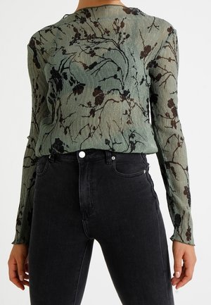 Femme portant un haut vert transparent à manches longues avec des motifs floraux noirs, rentré dans un jean noir taille haute.