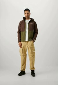 Καφέ και ελιά κομμάτι φερμουάρ με εφαρμοστή σχεδίαση, συνδυασμένο με μπεζ παντελόνι cargo με πολλές τσέπες. Μαύρα παπούτσια ολοκληρώνουν το σύνολο.