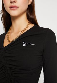 Sort langærmet bluse med samlede detaljer foran og hvidt broderet logo. Guldkæde med forlængede led.