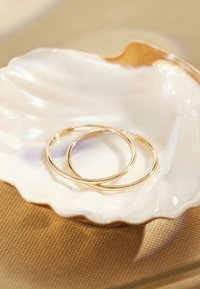 Gold hoop earrings resting in a scalloped, iridescent shell dish on a textured beige surface.