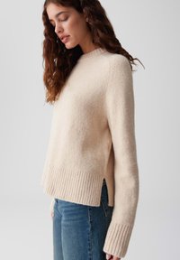 Donna con capelli ricci lunghi che indossa un maglione di lana color crema con polsini e orlo a costine, abbinato a jeans blu, in piedi di fronte a uno sfondo semplice.