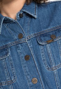 Jeansjacka med spetsig krage, två framfickor och mässingsknappar. Har blått tyg med kontrasterande orange sömmar och en logotagg.